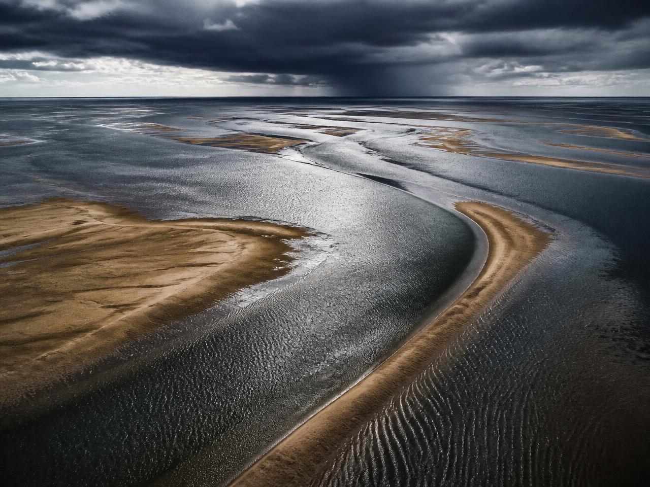 Tidal flats at low water with winding channels beneath a distant squall